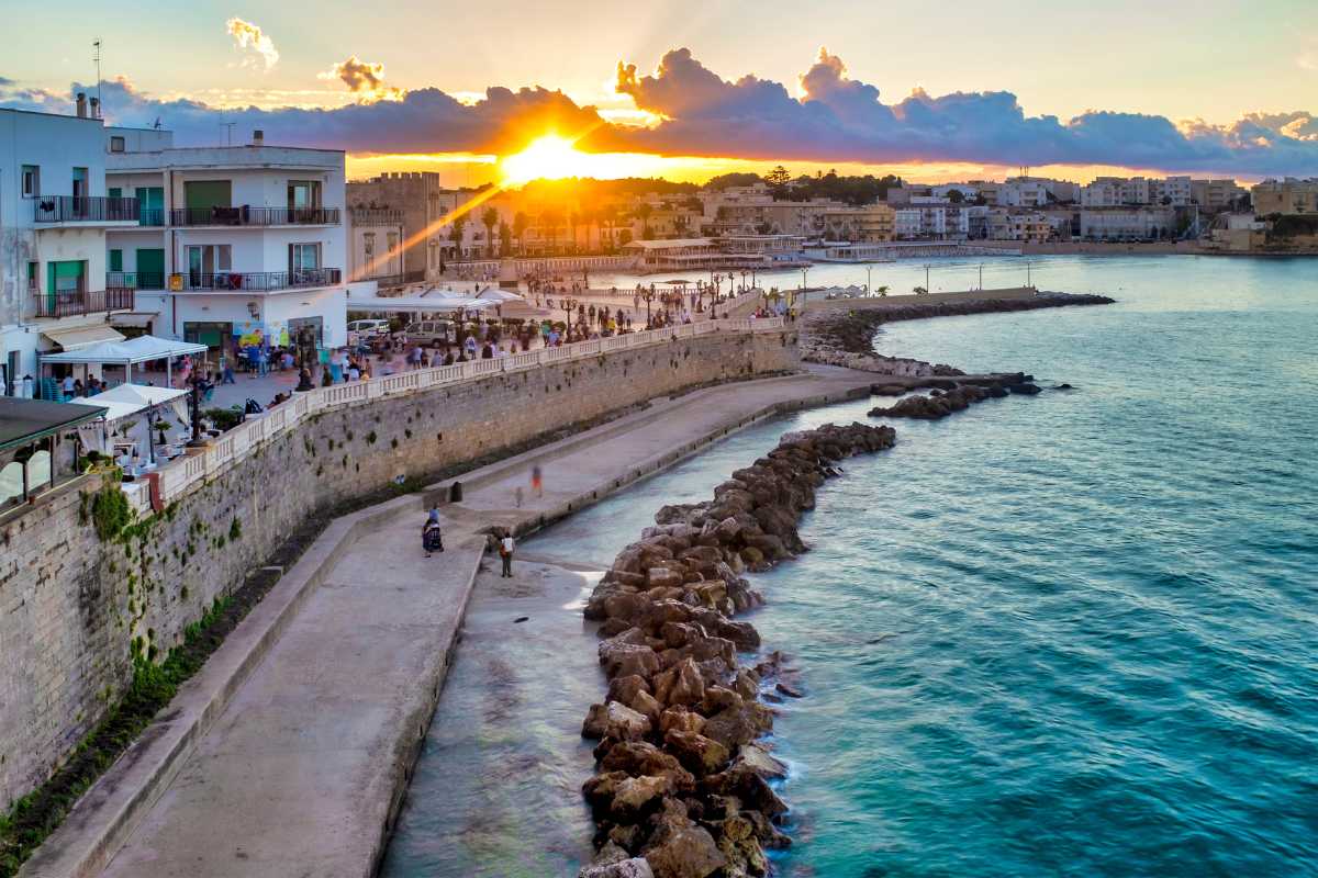 Vista panoramica di Otranto al tramonto, con il mare azzurro, il porticciolo e il borgo antico sullo sfondo. Atmosfera accogliente e vacanziera.