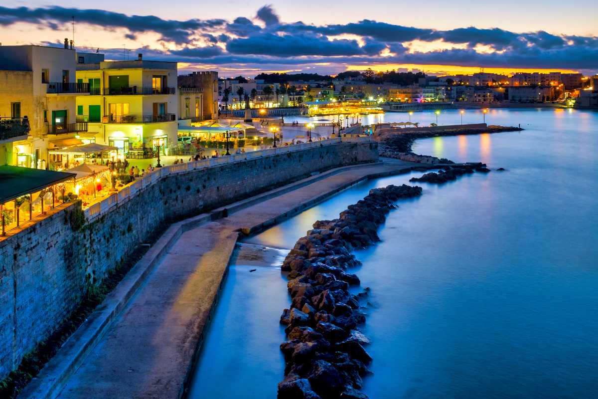 Viaggiatori in stazione ferroviaria di Lecce o Otranto, atmosfera rilassata, valigie colorate, treno regionale in arrivo, dettagli architettonici pugliesi